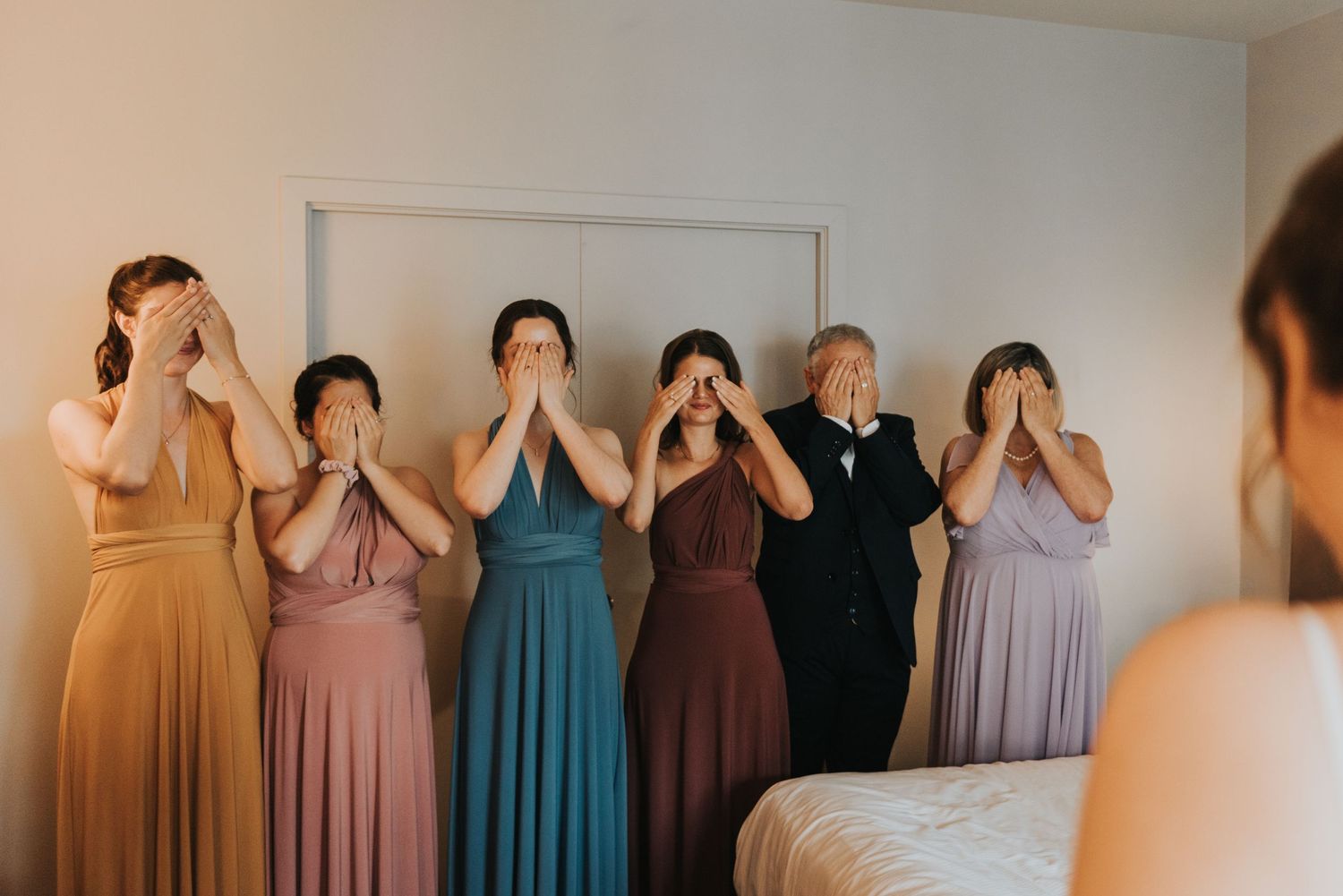 Wedding party members in different colored formal dresses cover their eyes during a playful reveal moment.