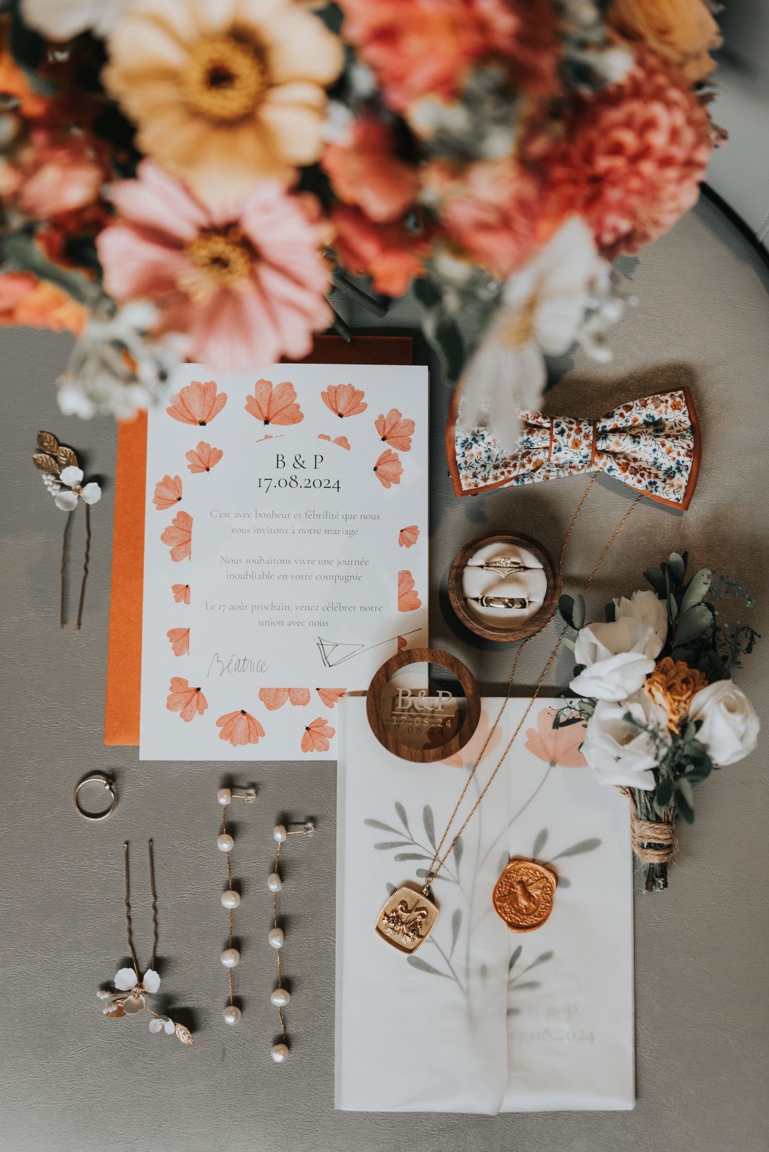 Wedding details flatlay with floral invitation, bow tie, rings, and jewelry against coral and peach flower backdrop.