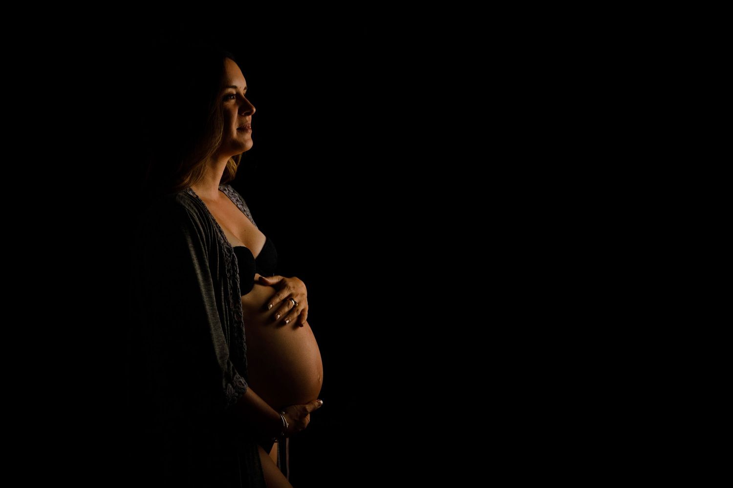 Silhouetted figure cradles pregnant belly against dark background in dramatic low-key maternity portrait.