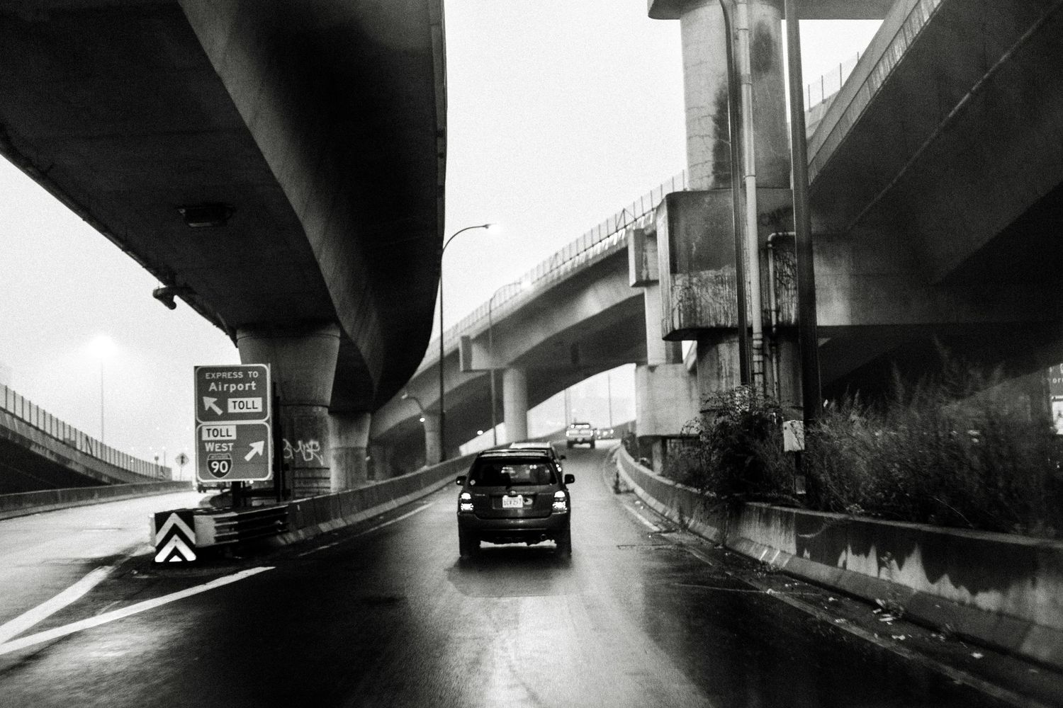 Vintage black and white photo of a car driving under elevated highway overpasses on a rainy day.