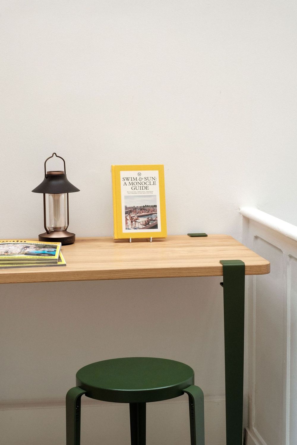Minimalist desk setup with wooden table, green stool, lantern and yellow book against white wall.