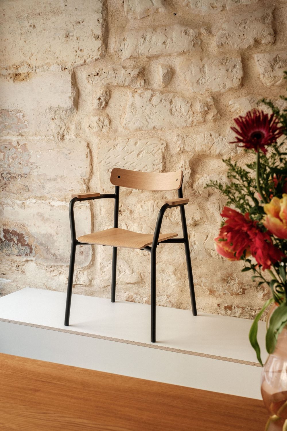 Modern wooden chair with black metal frame against textured stone wall with floral arrangement.