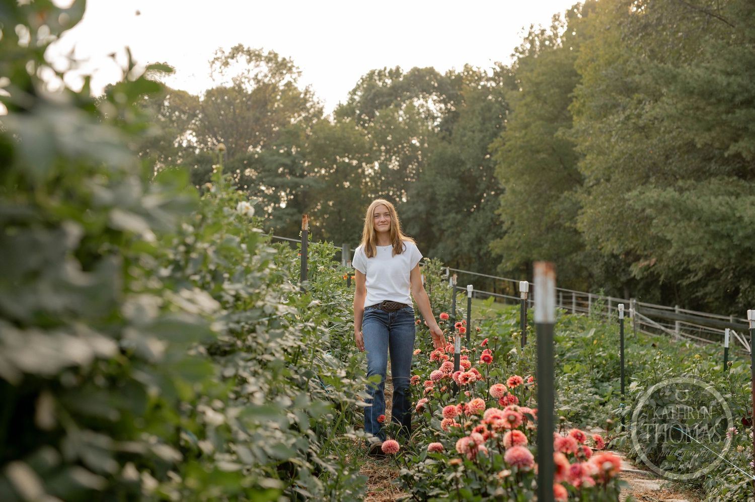 Woman in casual attire walks through garden path surrounded by blooming coral flowers and greenery.