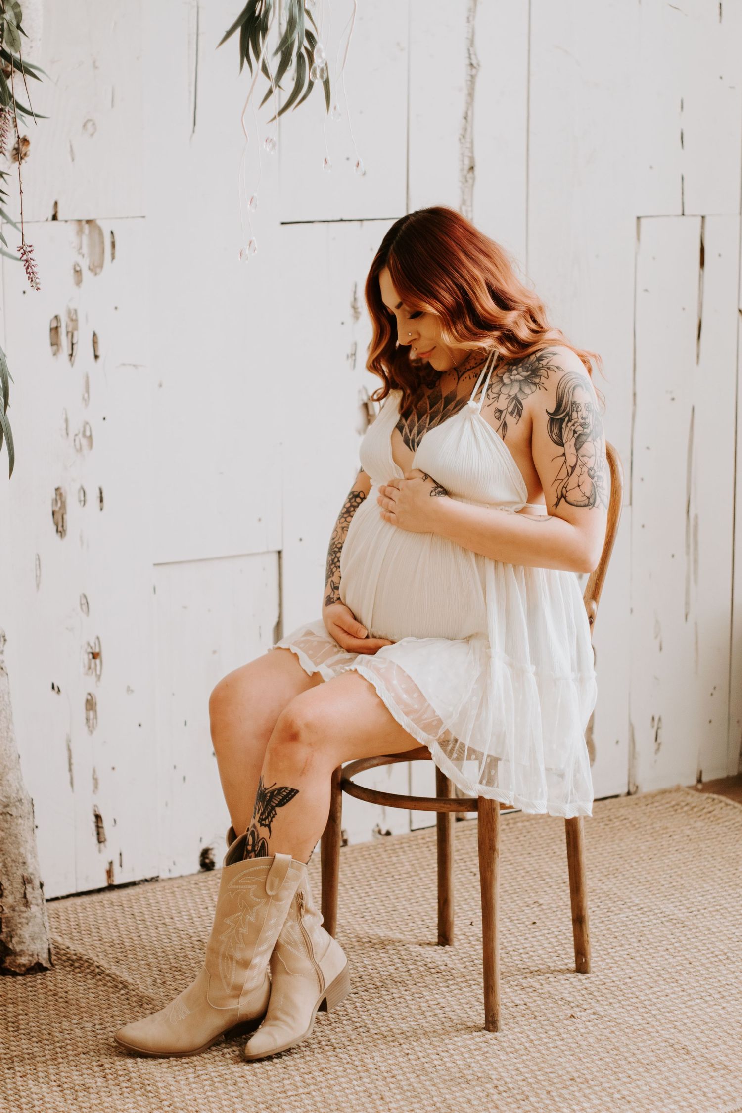 Pregnant woman in white dress and cowboy boots sits on wooden chair against rustic white wall with greenery.