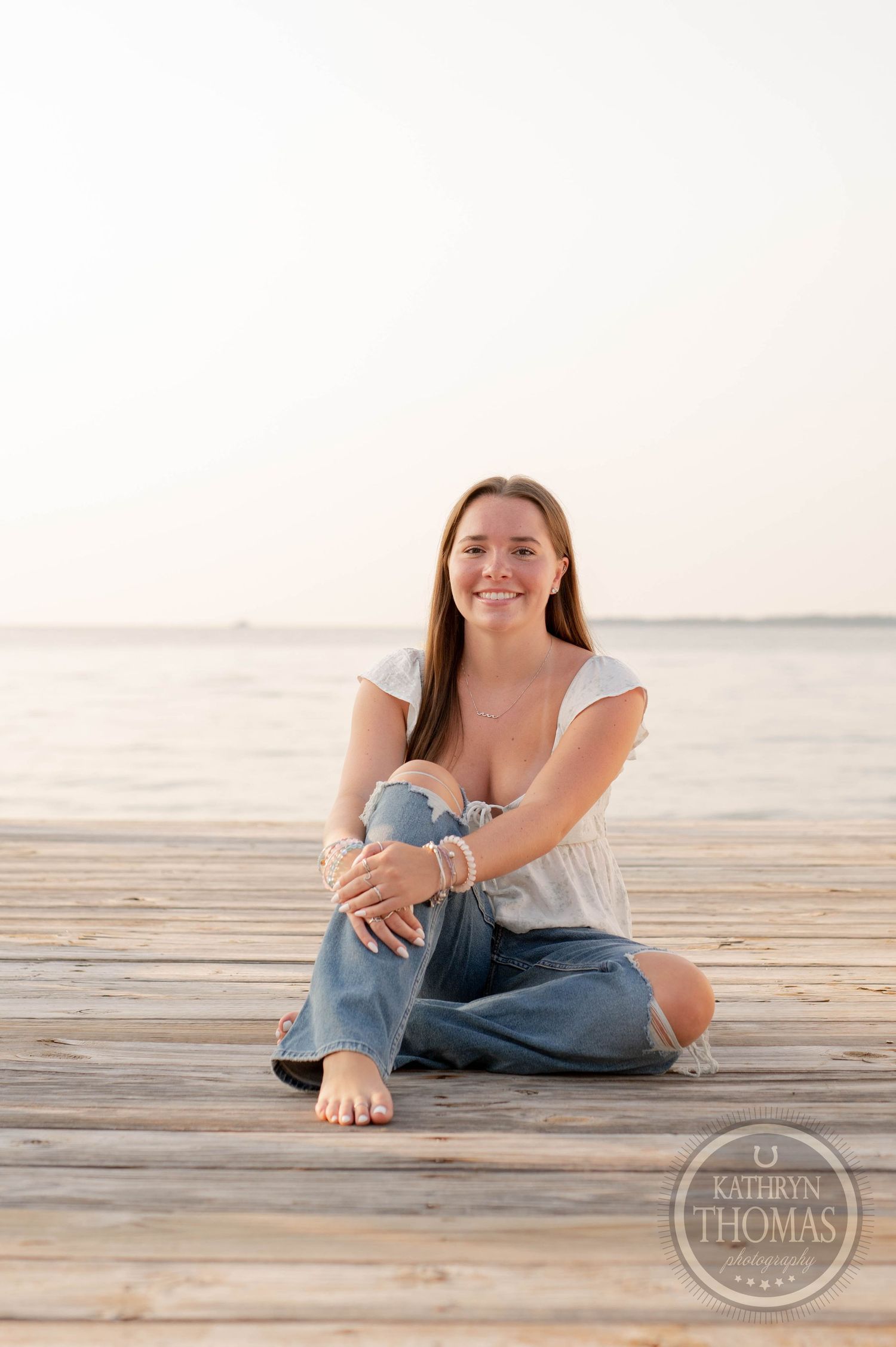 Person in casual attire sitting barefoot on wooden dock at sunset.
