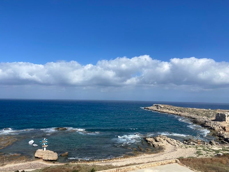 Rocky coastal landscape with turquoise waters and white clouds over a Mediterranean bay.