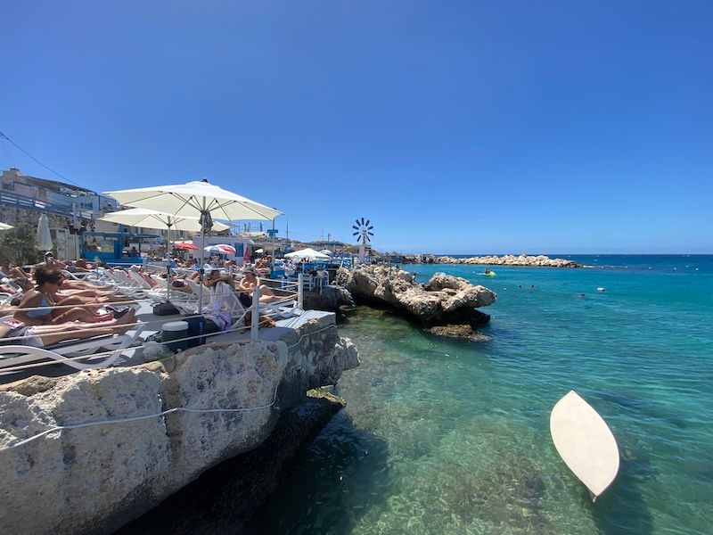 Beach umbrellas dot rocky coastal cliffs overlooking crystal clear turquoise Mediterranean waters on a sunny summer day.