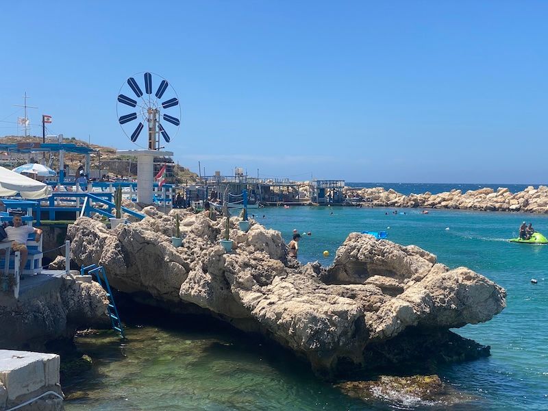 Blue windmill stands near rocky coastline with turquoise Mediterranean waters and coastal village in background.