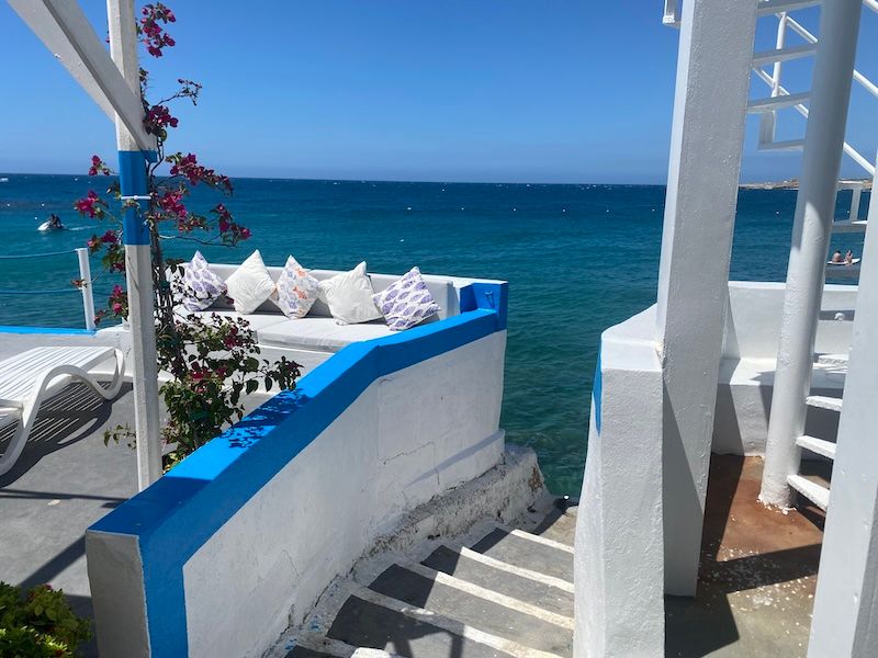 White and blue Mediterranean seaside terrace with lounge cushions overlooking turquoise ocean waters and flowering plants.