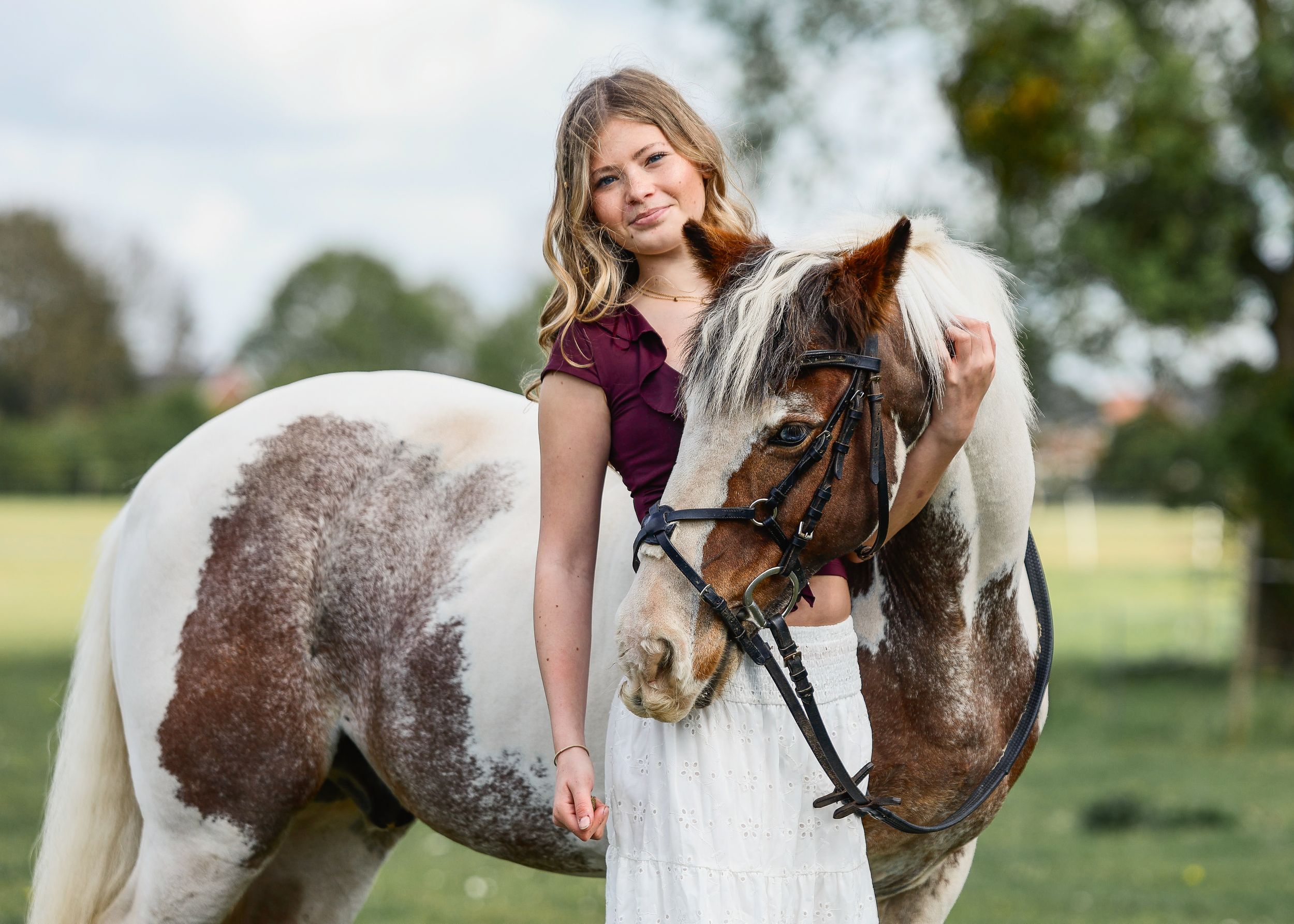 Young blonde girl in burgundy top and white skirt poses with a painted horse in a green field.
