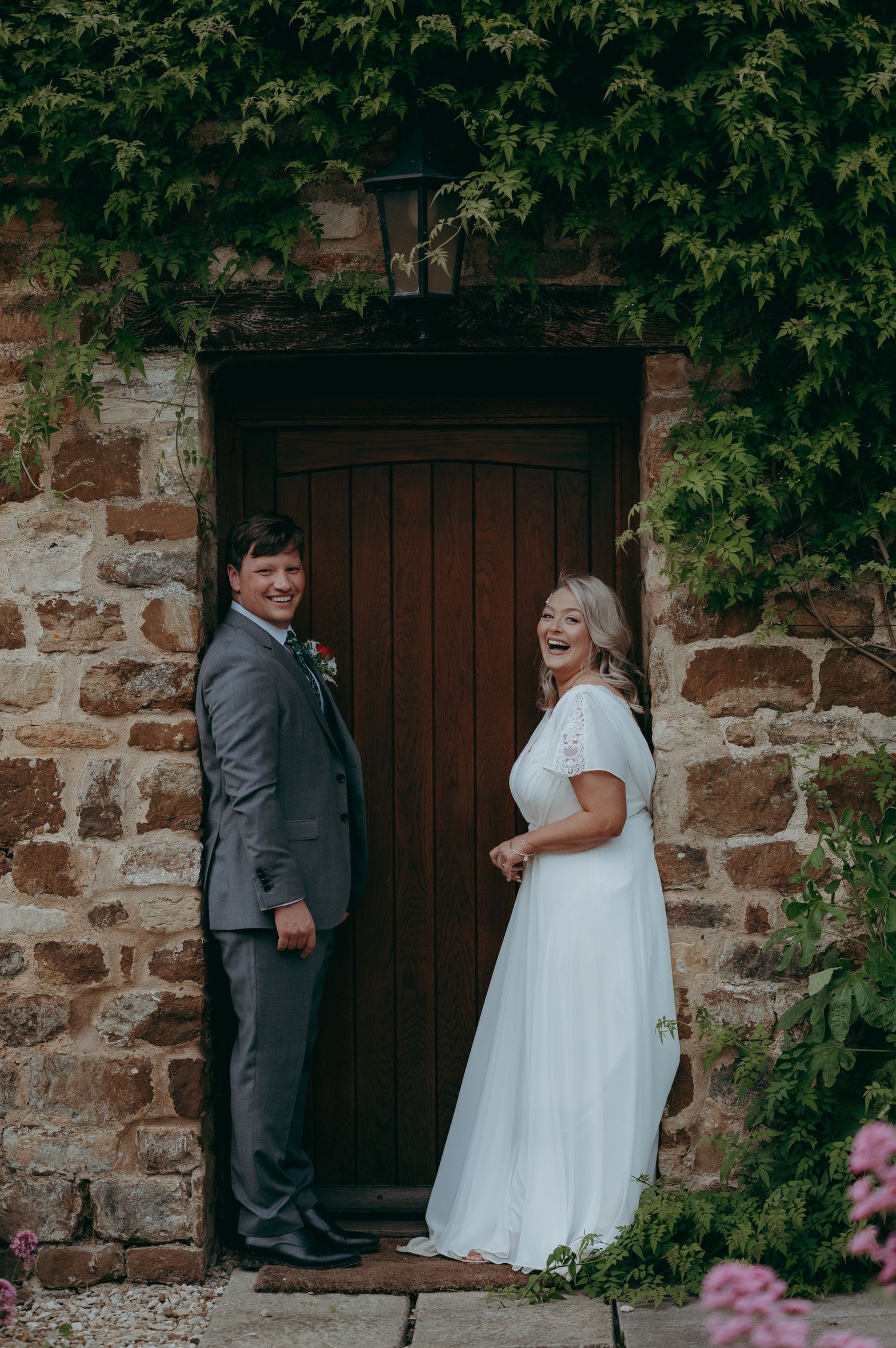 A rustic wedding photo featuring a couple posing by a stone wall and wooden door framed with green foliage.