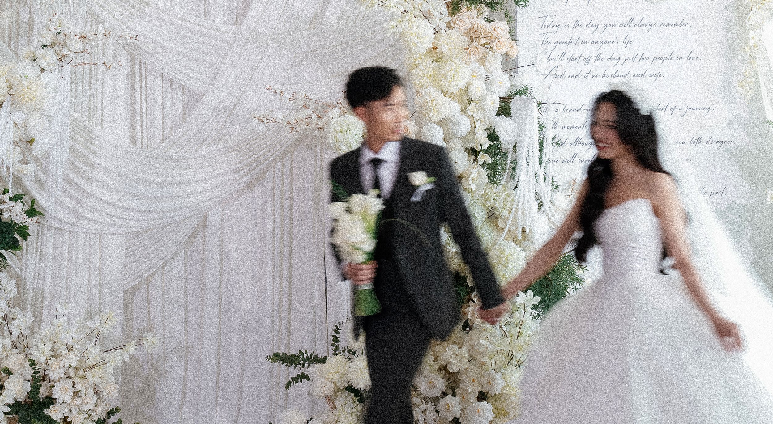 Couple holds hands at elegant white wedding ceremony surrounded by cascading floral arrangements and draped fabric.