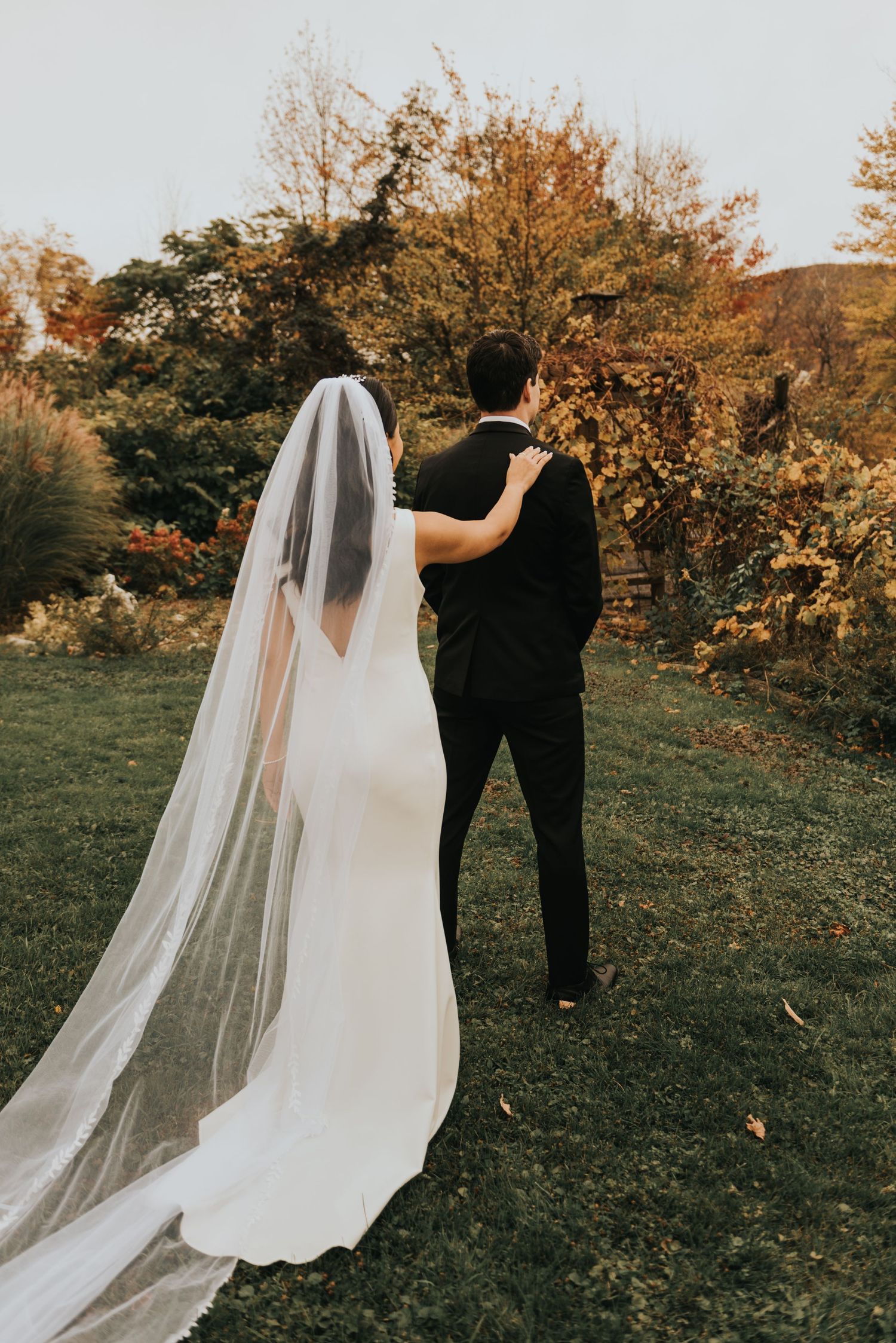 A couple in wedding attire walks on grass during autumn with a long flowing white veil trailing behind the bride.