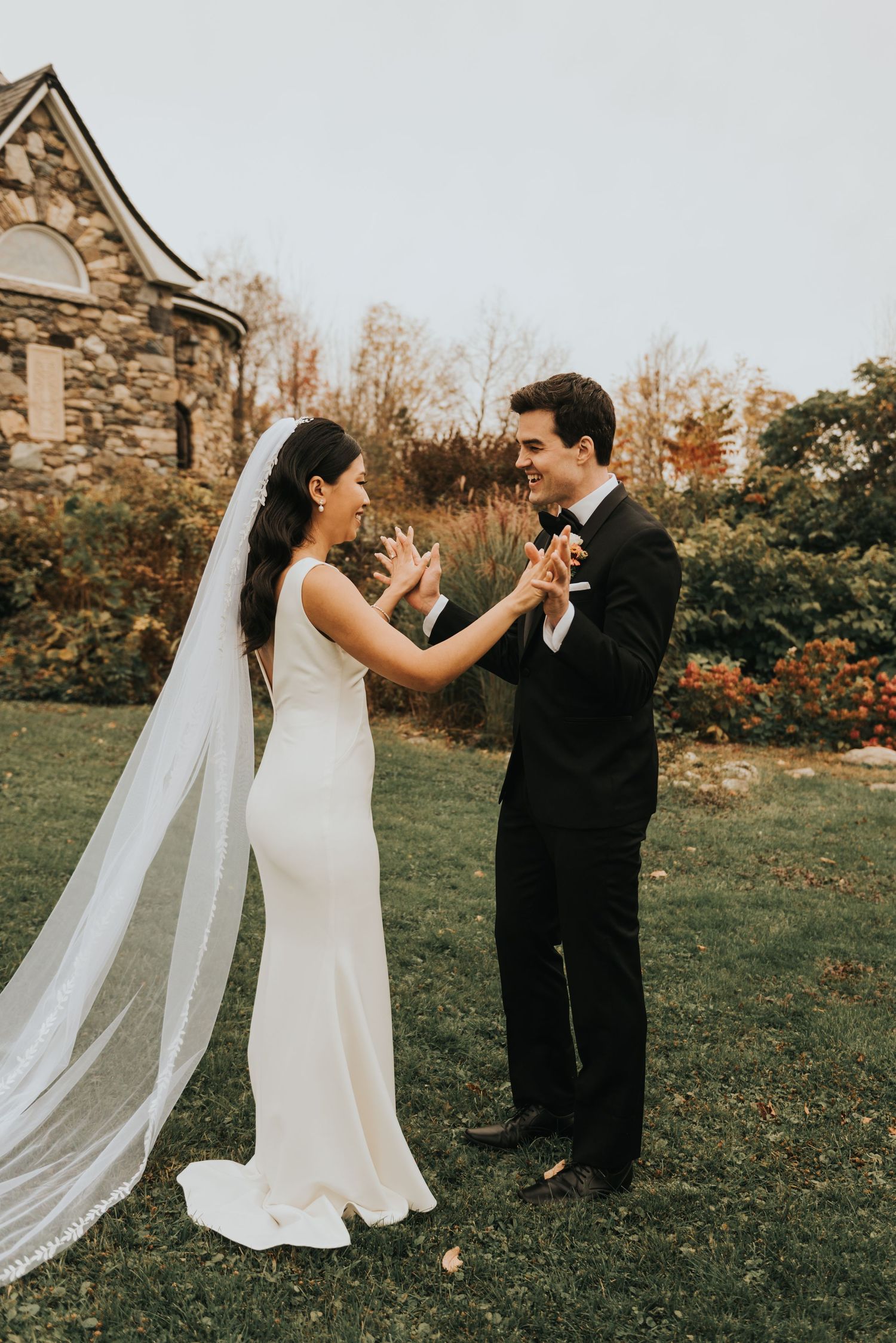 A bride in a sleek white gown and flowing veil dances with a groom in black tuxedo on grass near stone building.