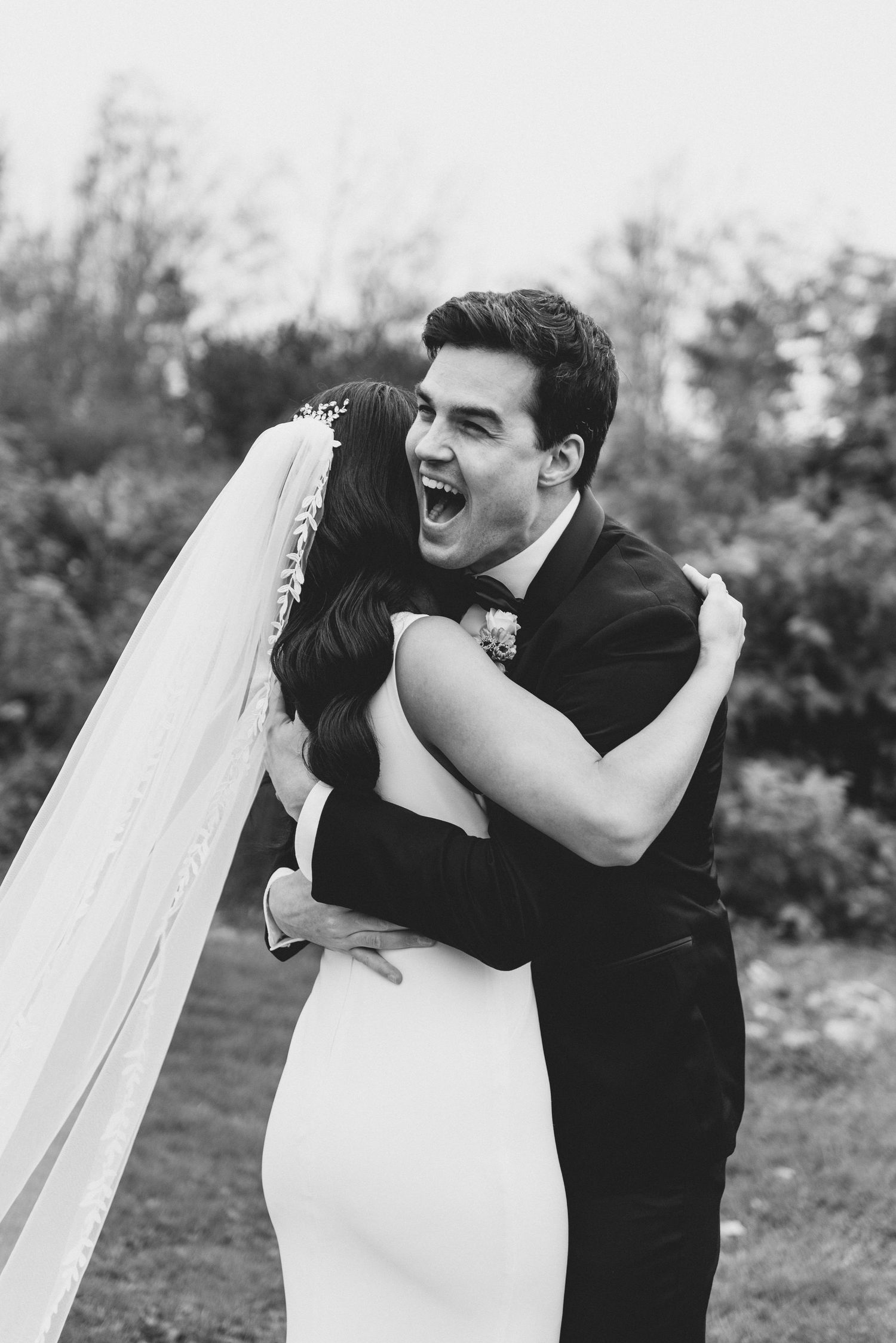 Black and white candid wedding photo showing joyful embrace between newlyweds outdoors with white veil flowing.