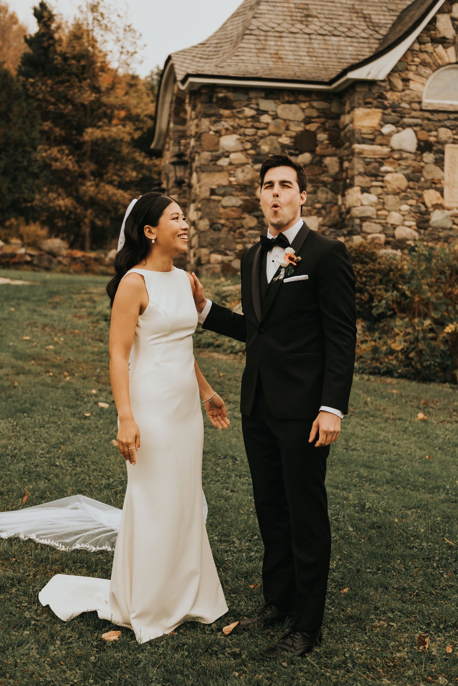 A couple shares a candid moment in front of a rustic stone building, wearing elegant wedding attire in a fall setting.