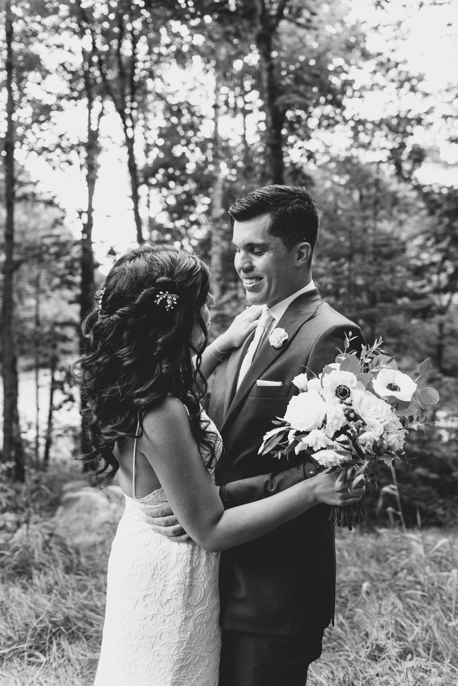 Black and white wedding photo of a couple in a forest setting with bride holding white floral bouquet.