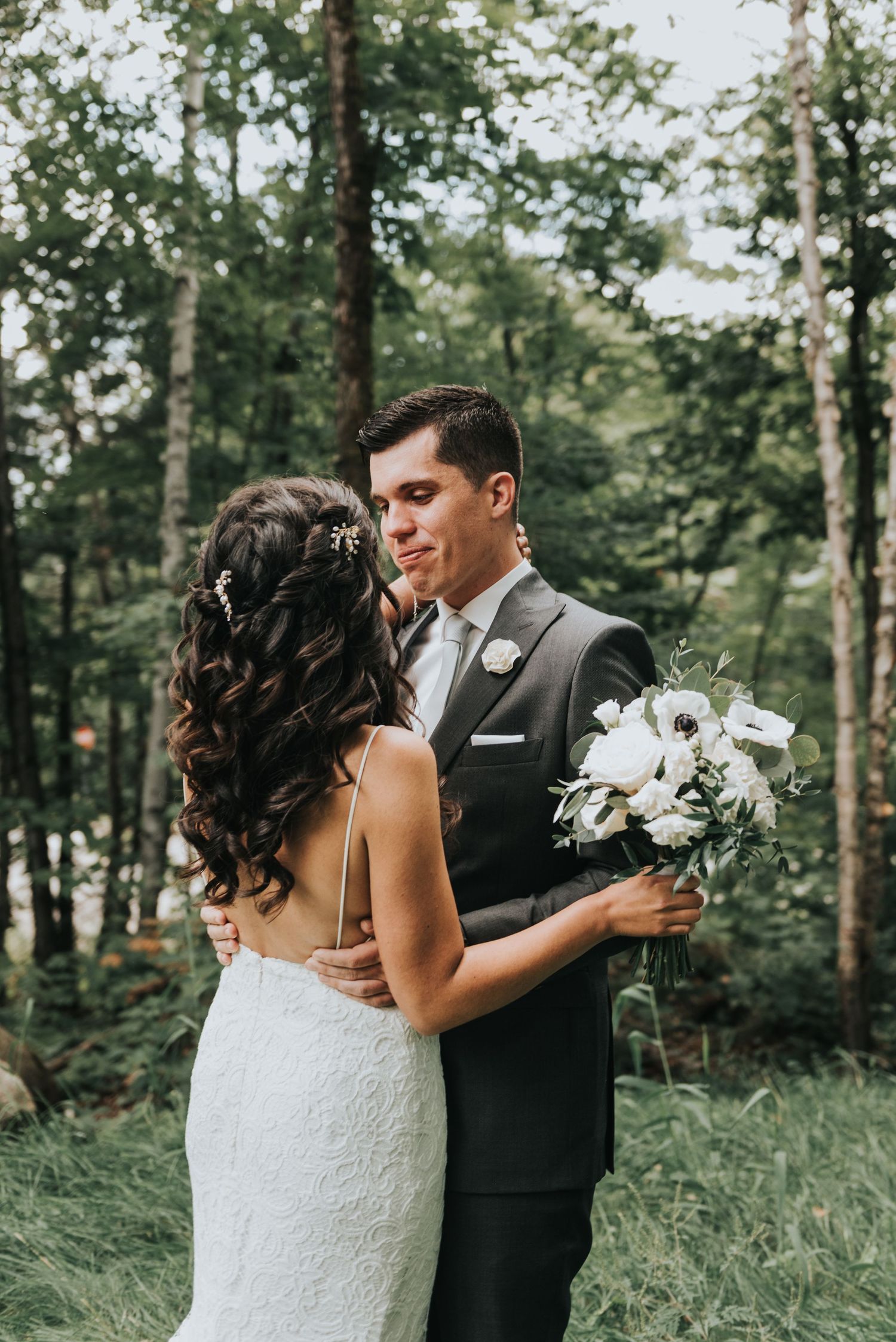 Newlywed couple stands intimately together in a forest setting with white bouquet and elegant wedding attire.