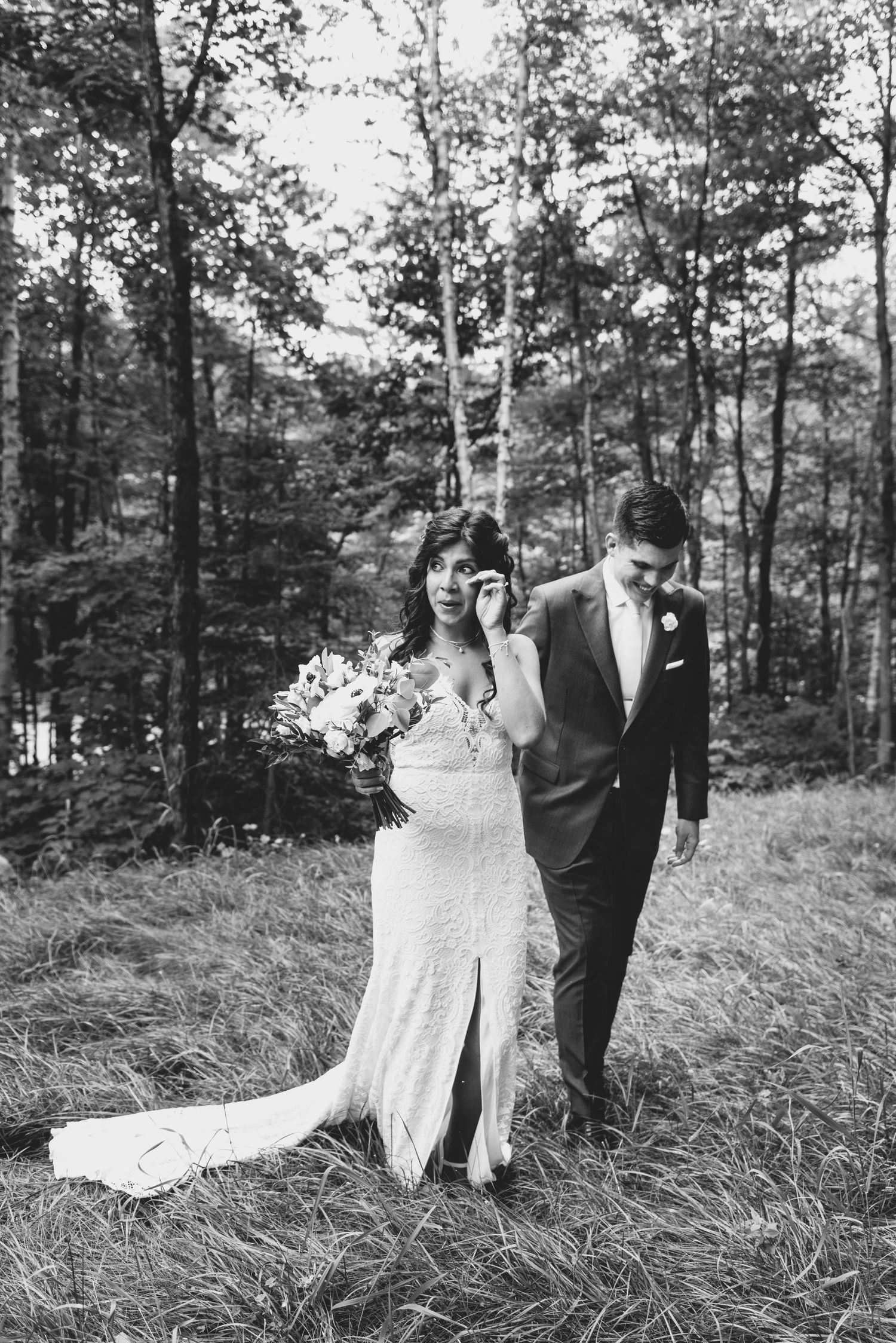 A black and white photo of a bride and groom walking through a wooded area at their outdoor wedding ceremony.