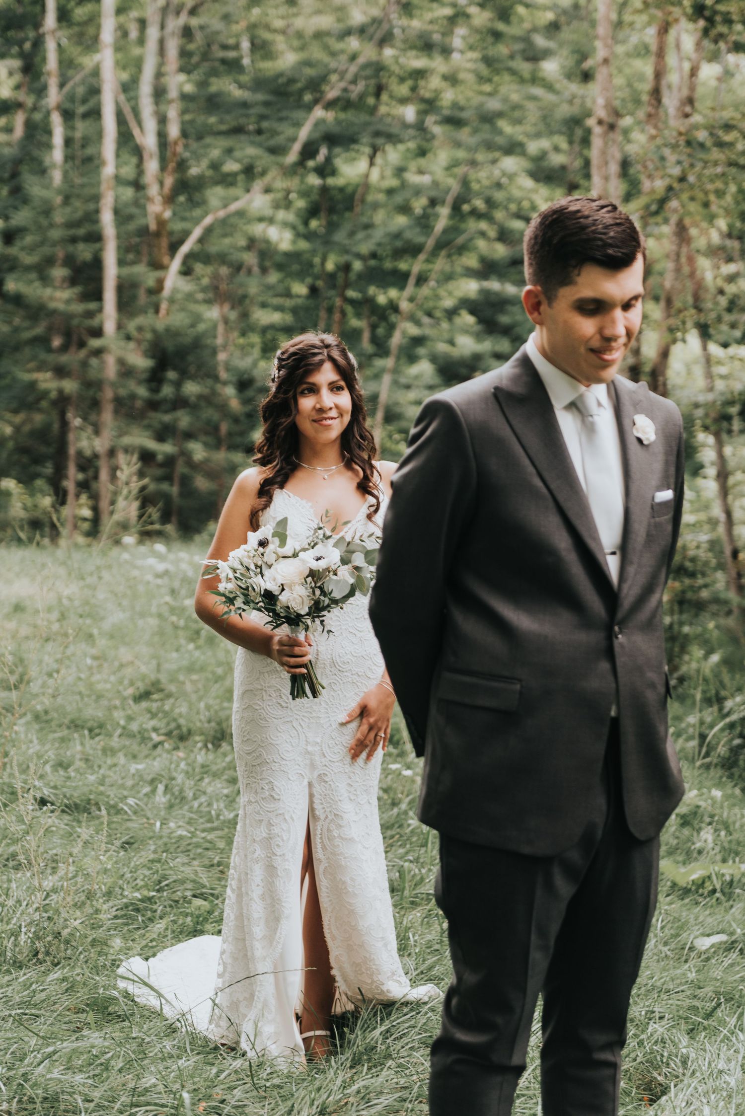 A newlywed couple poses in a wooded outdoor setting, with the bride holding a white floral bouquet and wearing a lace gown.