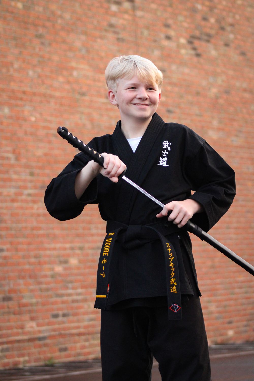 Young martial arts student poses with training sword while wearing traditional black uniform.