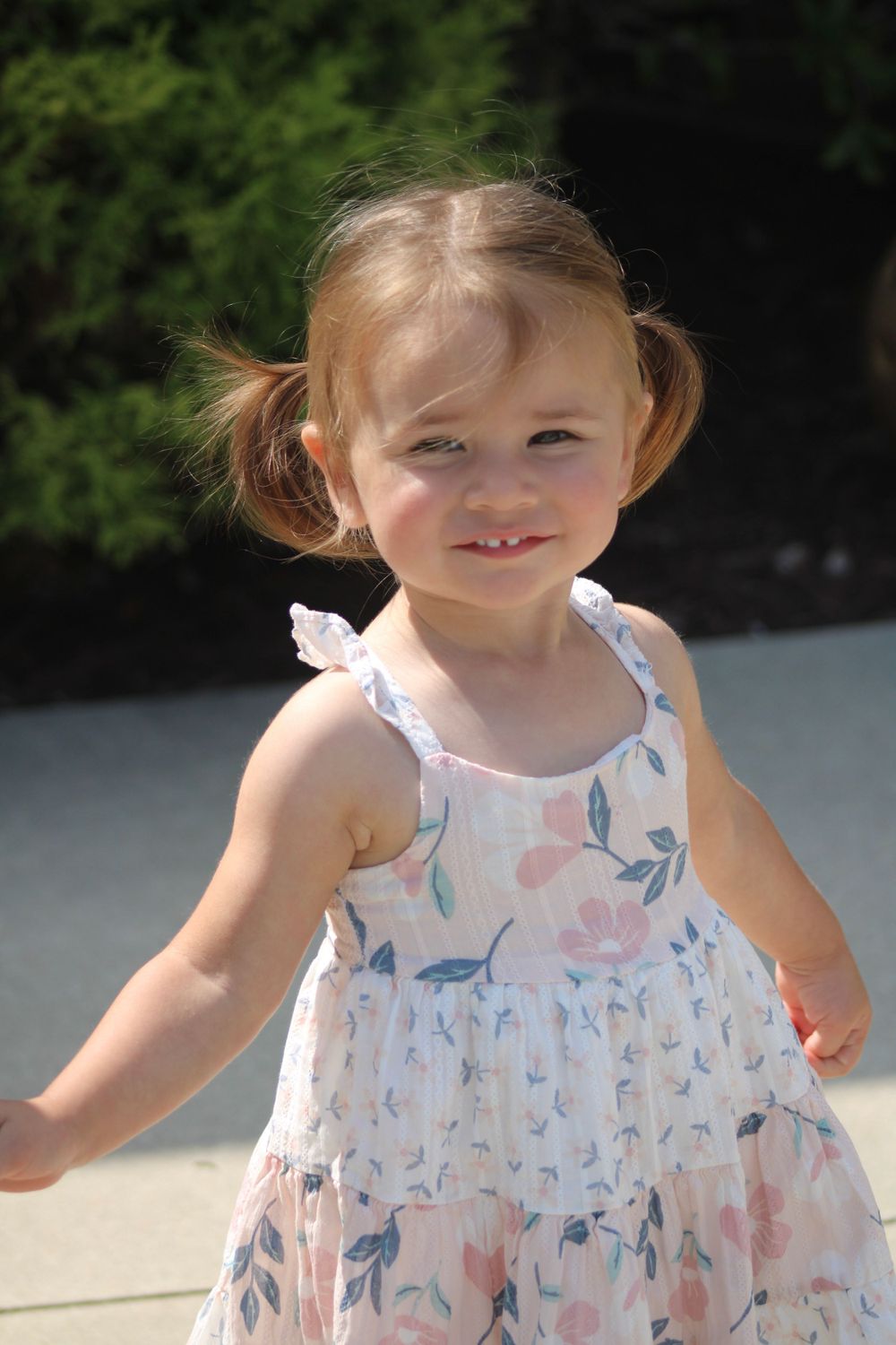 A young child in a floral summer dress smiles outdoors on a sunny day.