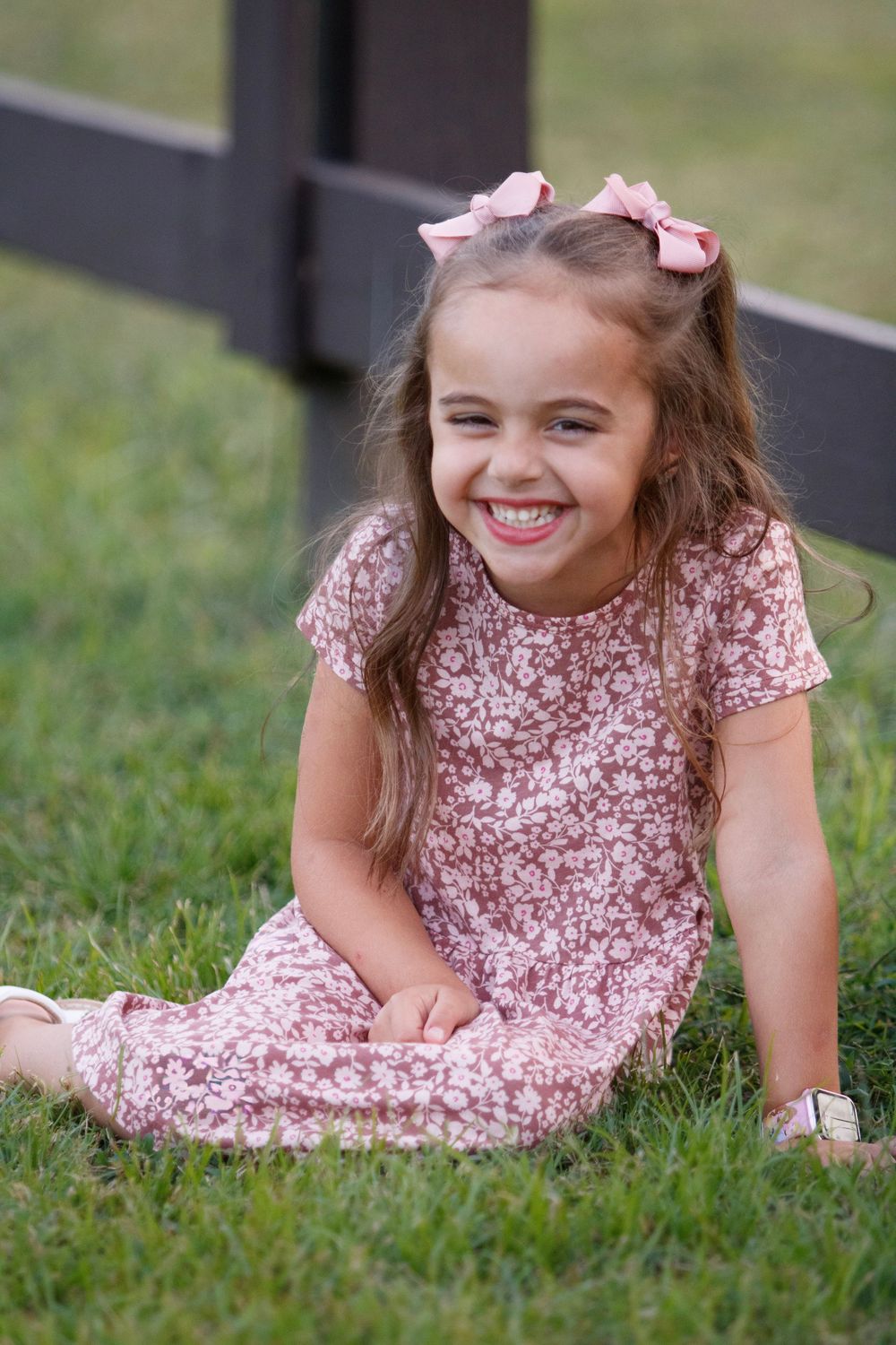 A child in a pink floral dress lies on green grass while smiling and playing outdoors.