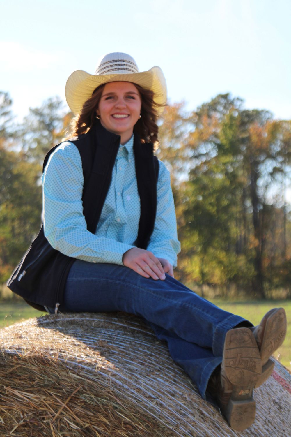 Person in western wear relaxes on hay bale wearing cowboy boots and hat.