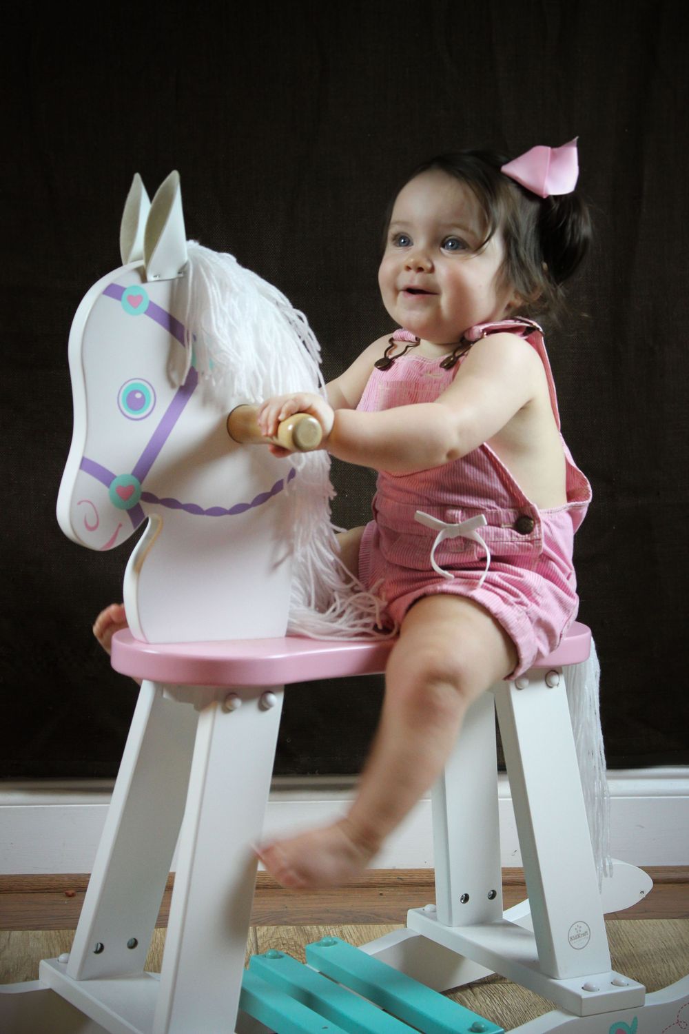 A child plays on a white rocking horse toy wearing pink clothing against a dark background.