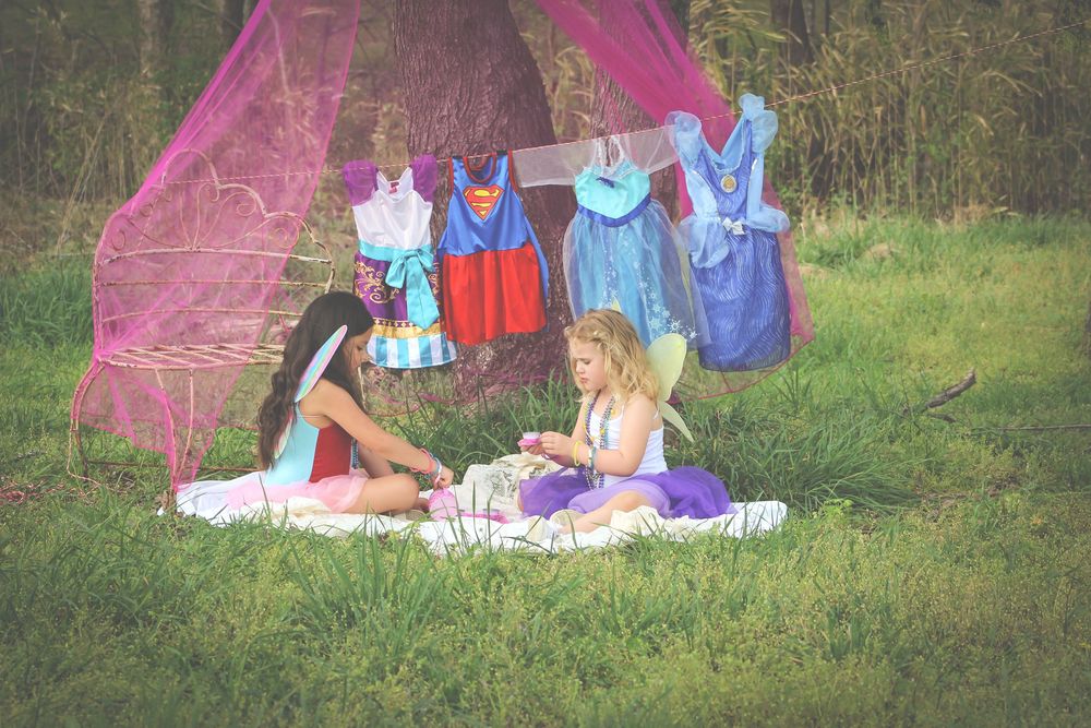 A pink umbrella sits among scattered clothing and blankets on a grassy lawn in a garden setting.
