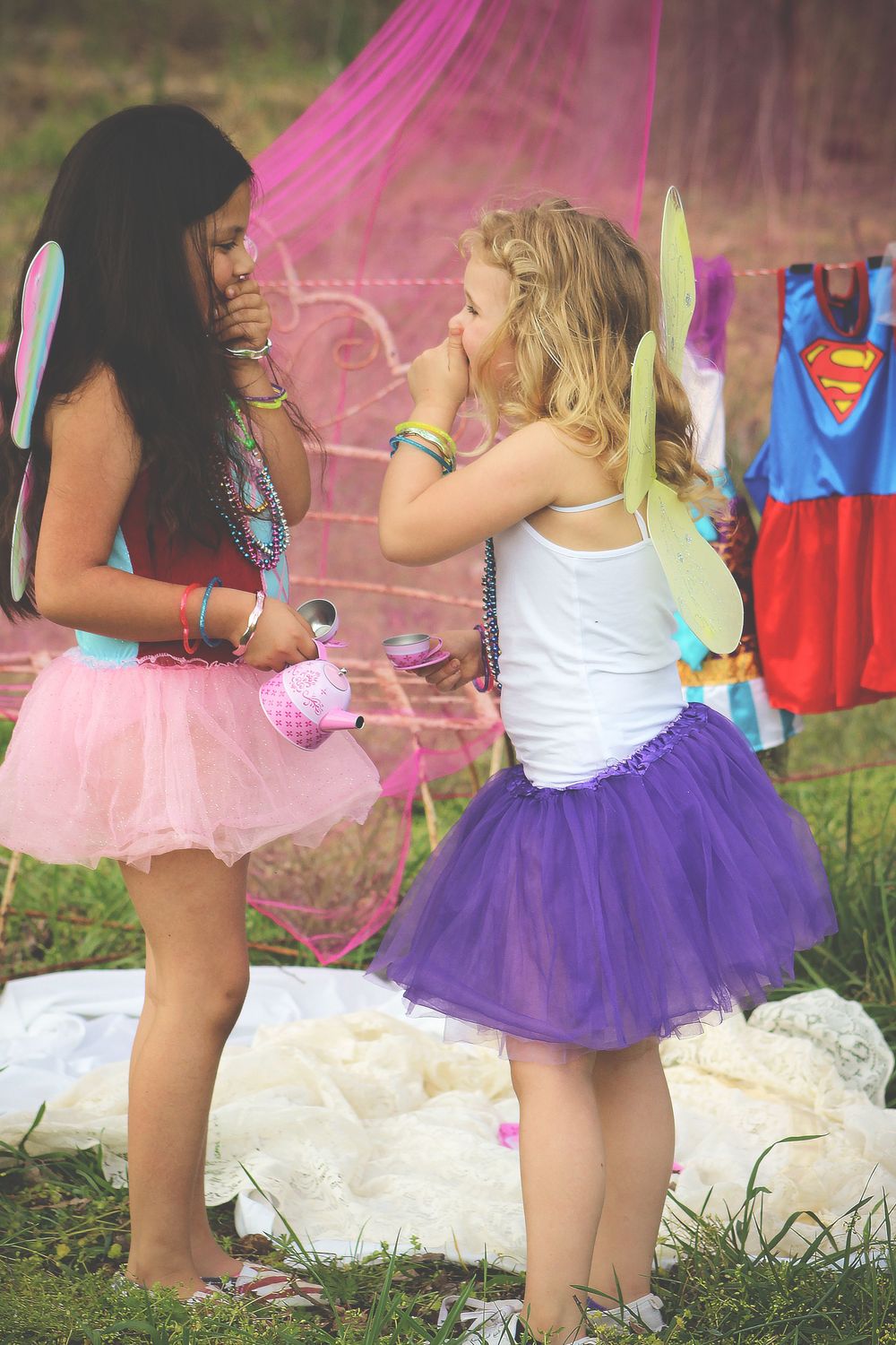 Two friends in pink and purple tutus and fairy wings share a playful moment outdoors with superhero costume in background.