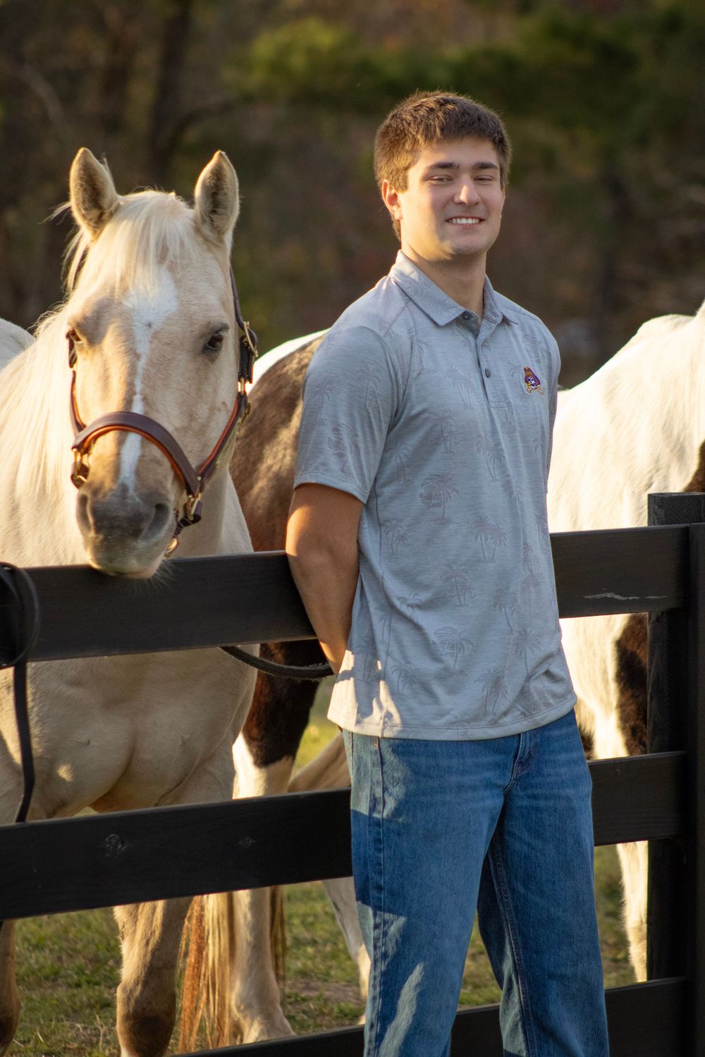 Young man in gray polo shirt standing next to white horse behind black fence.
