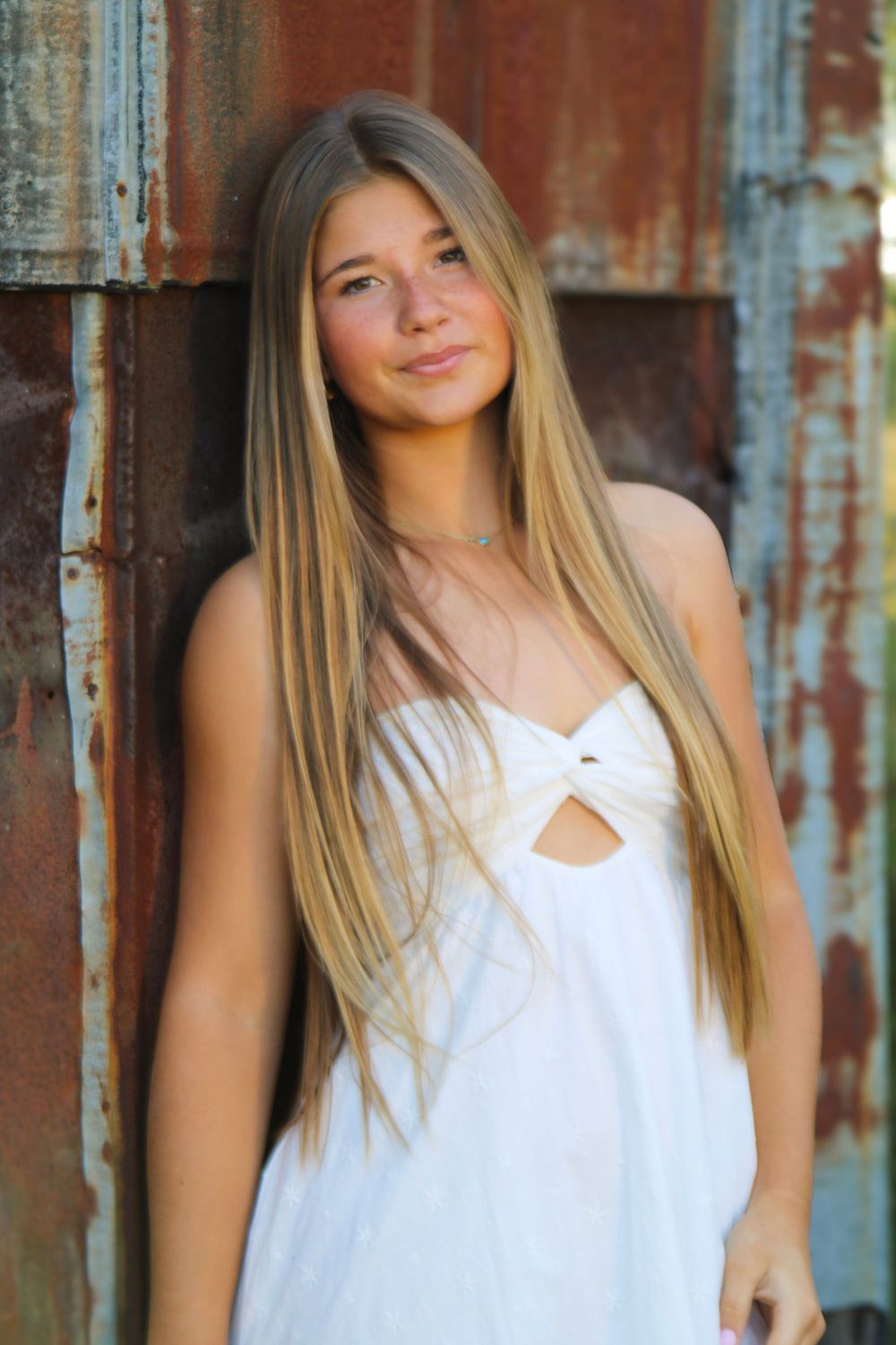 Portrait against rustic metal wall wearing white summer dress with long blonde hair.