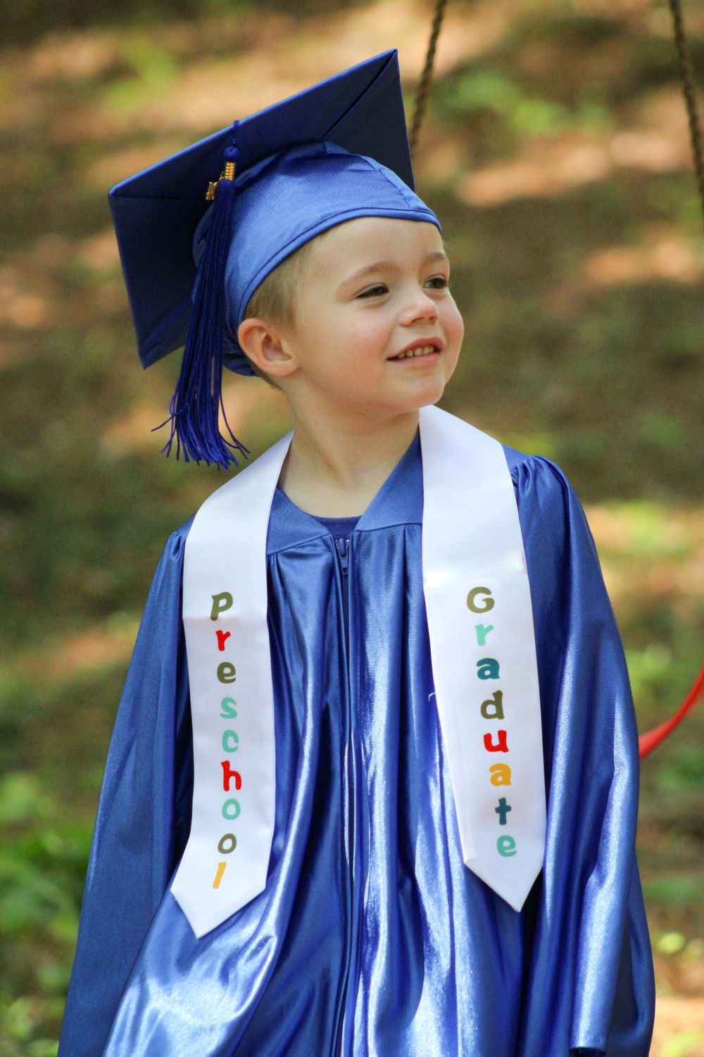 A smiling preschool graduate wearing a blue cap and gown with white sash outdoors.