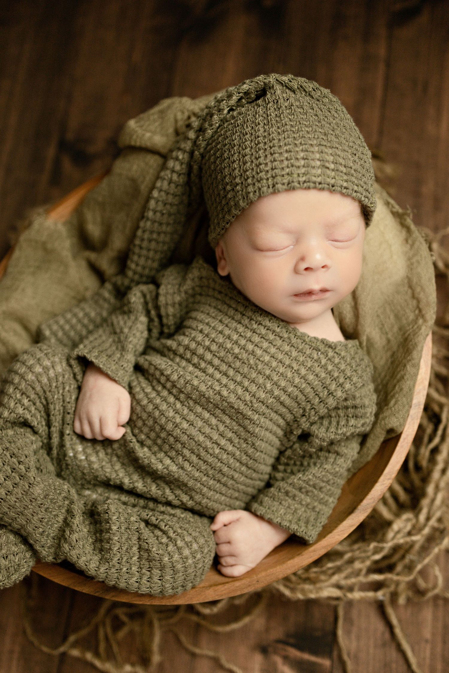 Sleeping baby wearing cozy olive green knit sweater and matching hat rests peacefully in wooden basket.