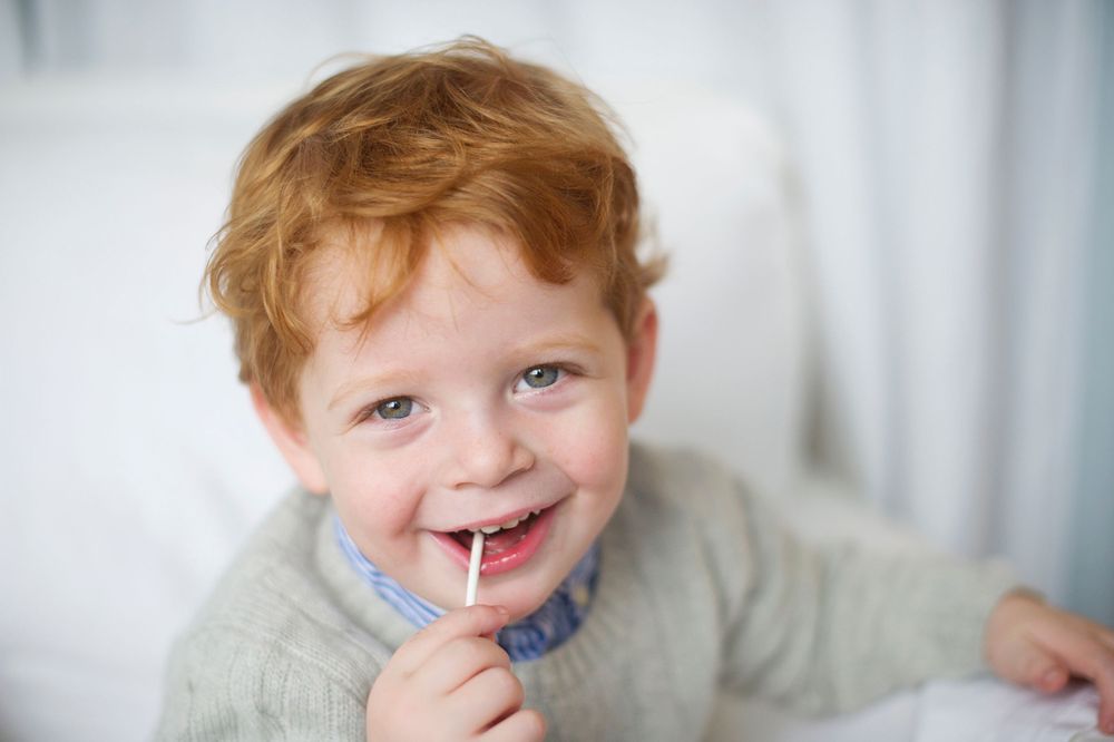 Smiling toddler with red hair enjoying a lollipop, captured by Phoebe Lee Photo during a family session in Toronto