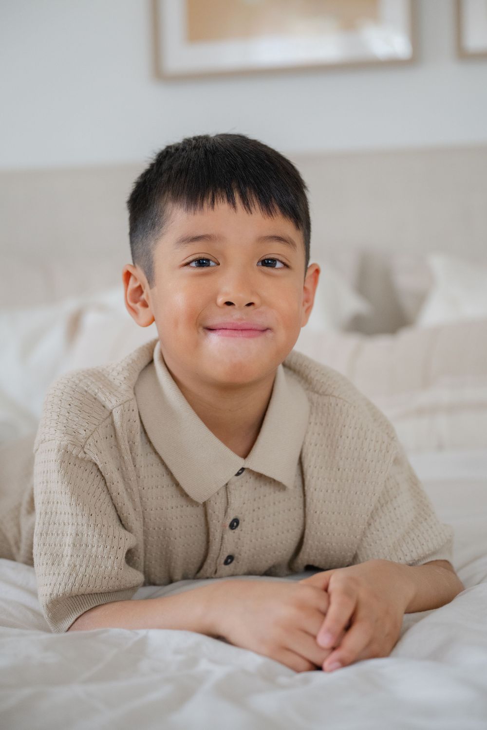 A young child in a beige knit sweater lies on white bedding with a playful expression. Captured by Phoebe Lee Photo of Toronto, Ontario.