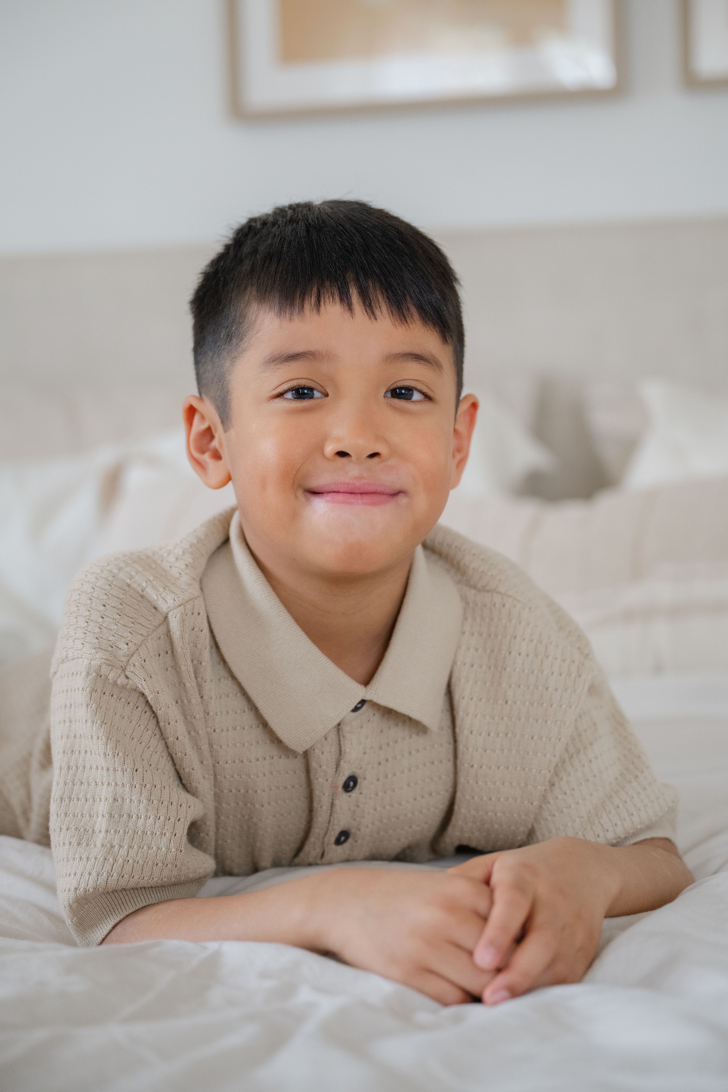 A young child in a beige knit sweater lies on white bedding with a playful expression. Captured by Phoebe Lee Photo of Toronto, Ontario.