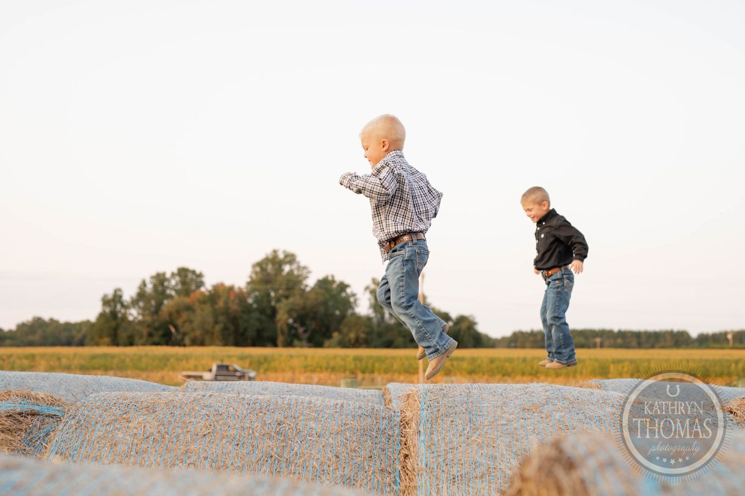 Children jumping between round hay bales in a field during sunset.
