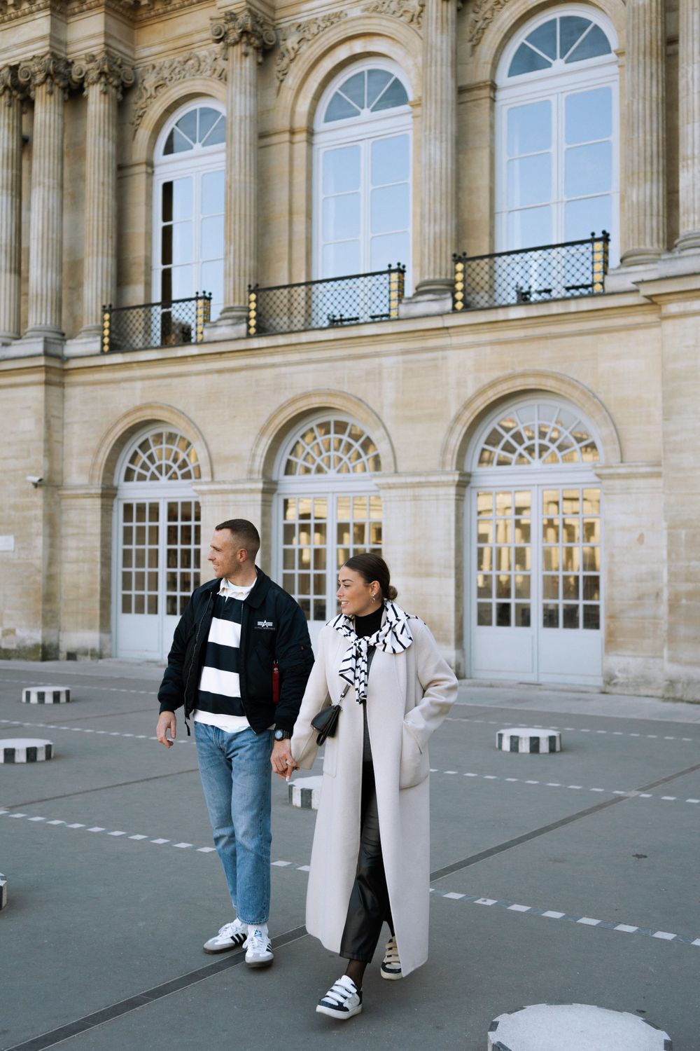 Two people stroll past elegant classical architecture with arched windows and stone columns at Palais Royal in Paris.