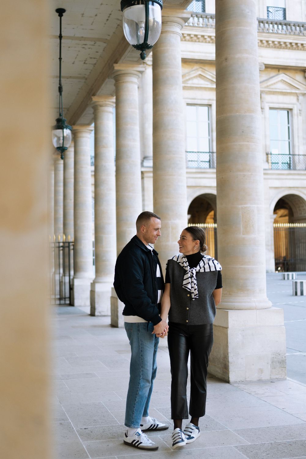 A couple shares a romantic moment while standing among elegant stone columns in a classical European building's arcade.