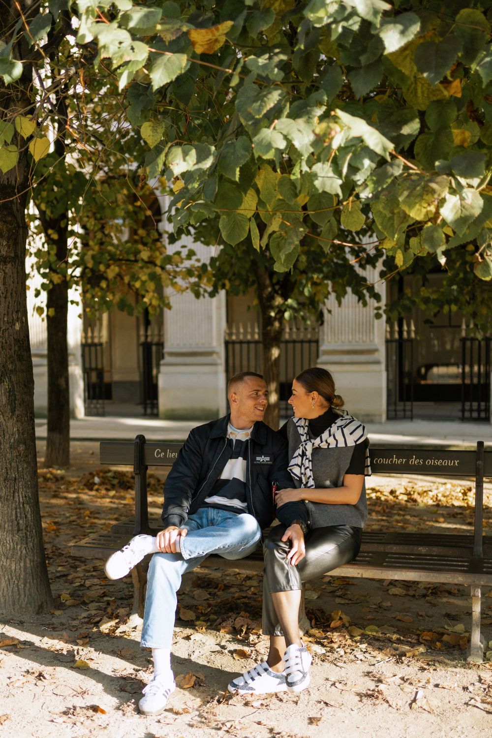 A couple sits together on a wooden park bench under autumn trees, sharing an intimate moment.
