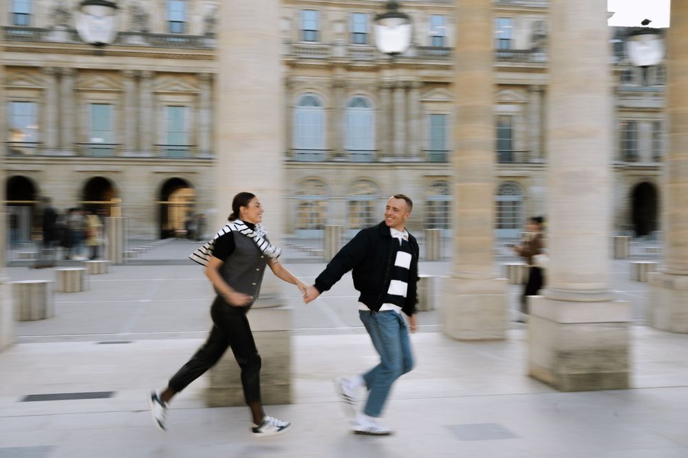 Two people playfully running between columns in front of an ornate European building with classical architecture.
