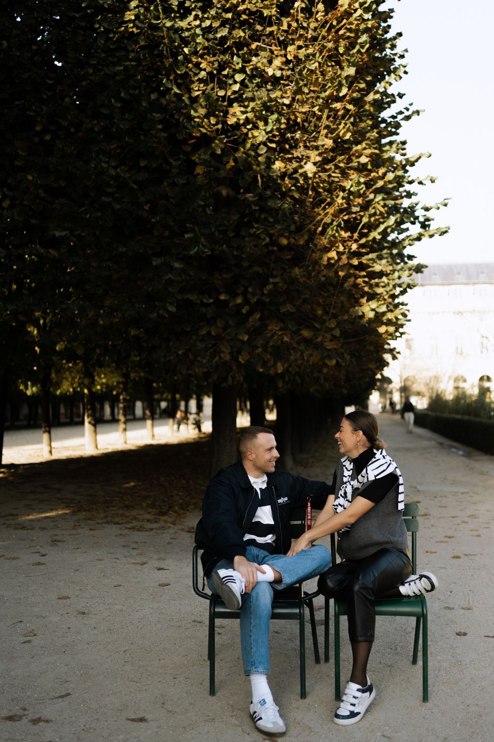 Two people sitting on green chairs having a conversation along a tree-lined path in a park on an autumn day.