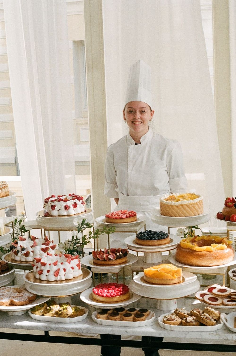 A pastry chef stands behind an elegant display of various cakes and desserts including fruit tarts and cream cakes.