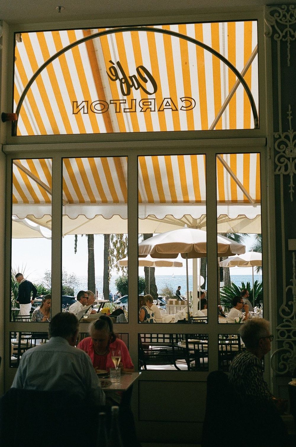 View through window of Cafe Carlton's outdoor terrace with orange and white striped awning and diners seated at tables.
