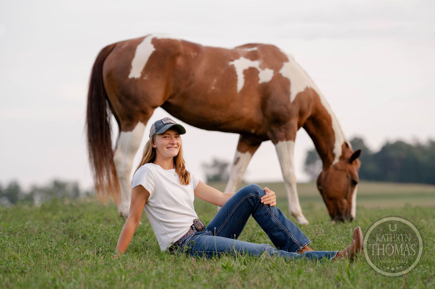 Paint horse feeds on grass while companion in casual wear relaxes in summer field.