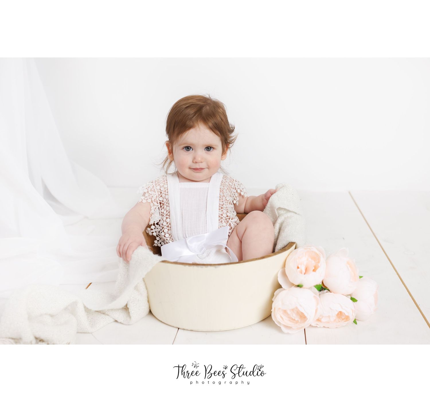 A small child sits in a cream-colored metal basin with soft pink roses and white fabric nearby for a portrait session.