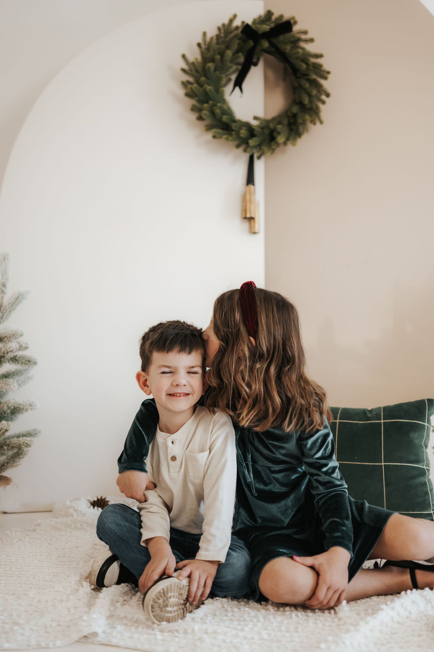 Two people sitting on a fluffy white carpet under a green holiday wreath hanging on a cream colored wall.