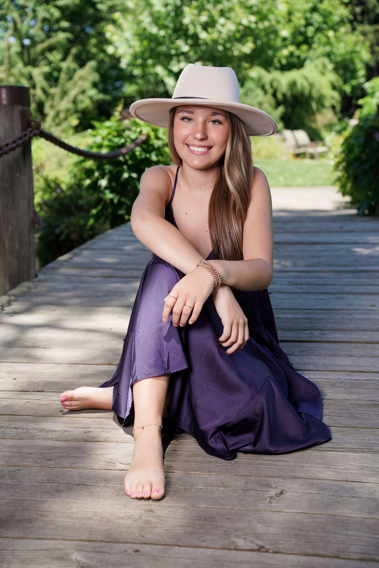 Woman in purple dress and tan hat sitting casually on wooden boardwalk.